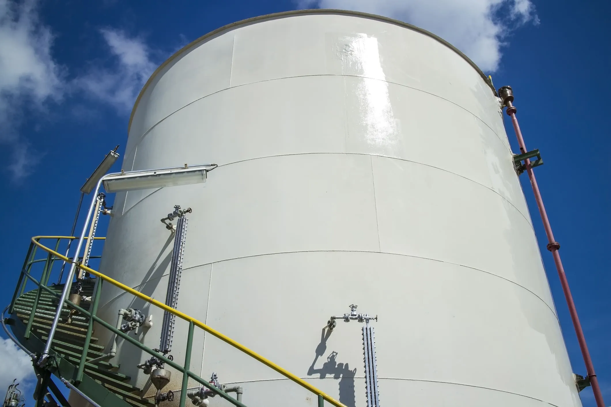 Large white protective-coated industrial storage tank with access stairs and yellow safety railings against a blue sky