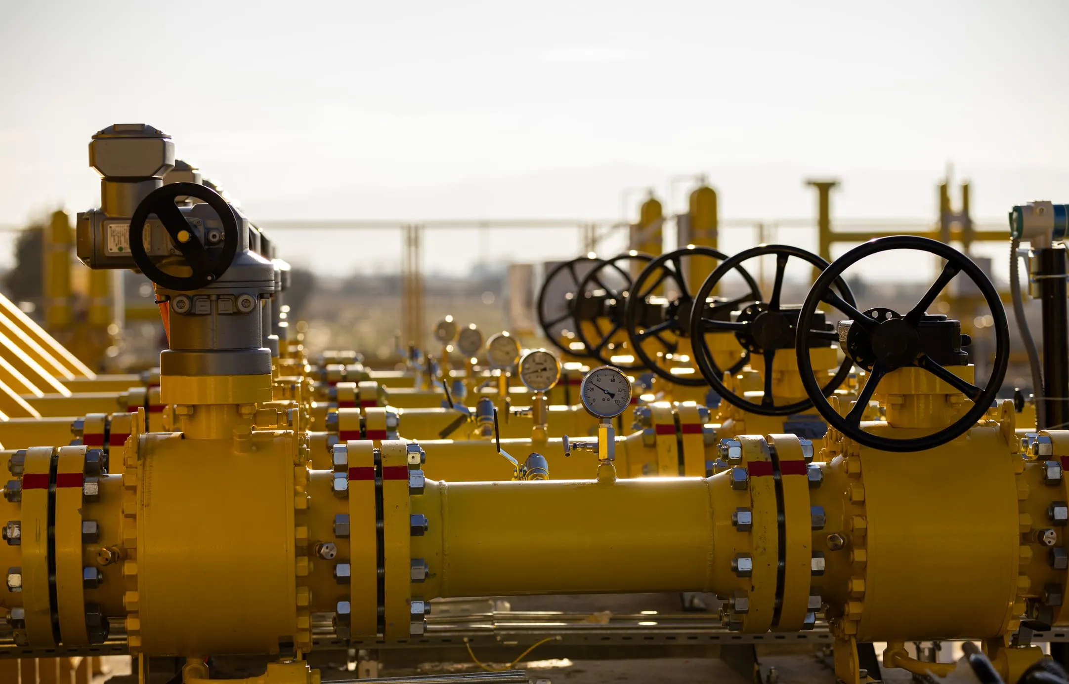 Coated yellow pipeline manifold with valves, hand wheels, and pressure gauges at an industrial facility in golden light