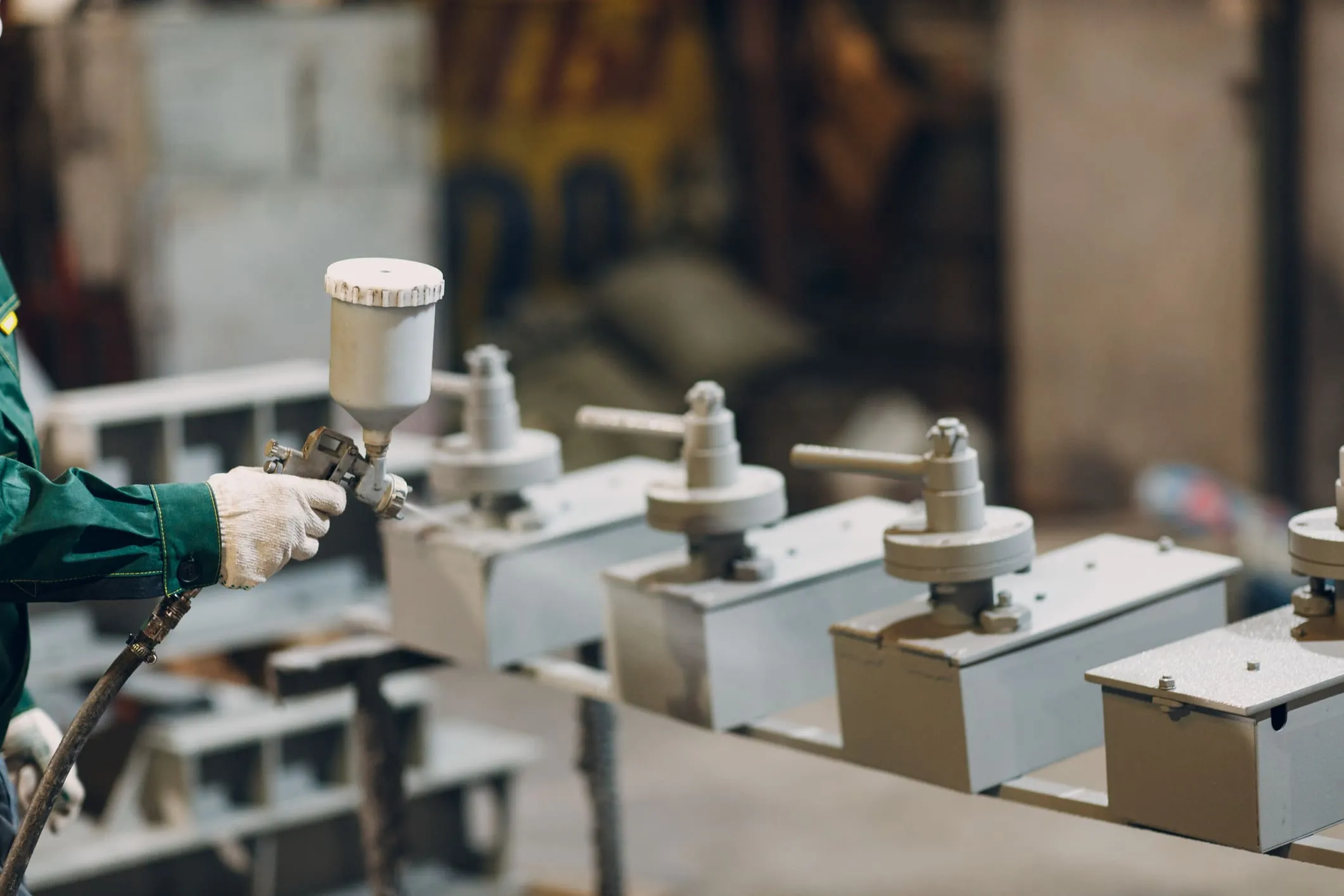 Gloved applicator using an HVLP spray gun to coat a row of fabricated steel parts on a shop line